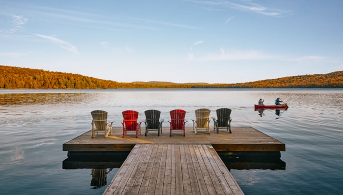 A row of six Muskoka chairs on a dock face a calm lake with canoe and fall colour in the distance.