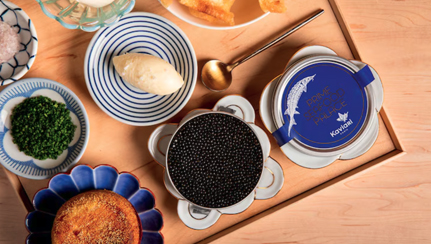 Plateau gastronomique comprenant du caviar, des herbes hachées, une tartinade crémeuse et une cuillère dorée, placé sur une surface en bois.