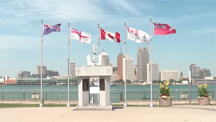 Le monument naval de Windsor, en Ontario, avec une sculpture devant six drapeaux flottants et une ville surplombant l’eau en toile de fond.