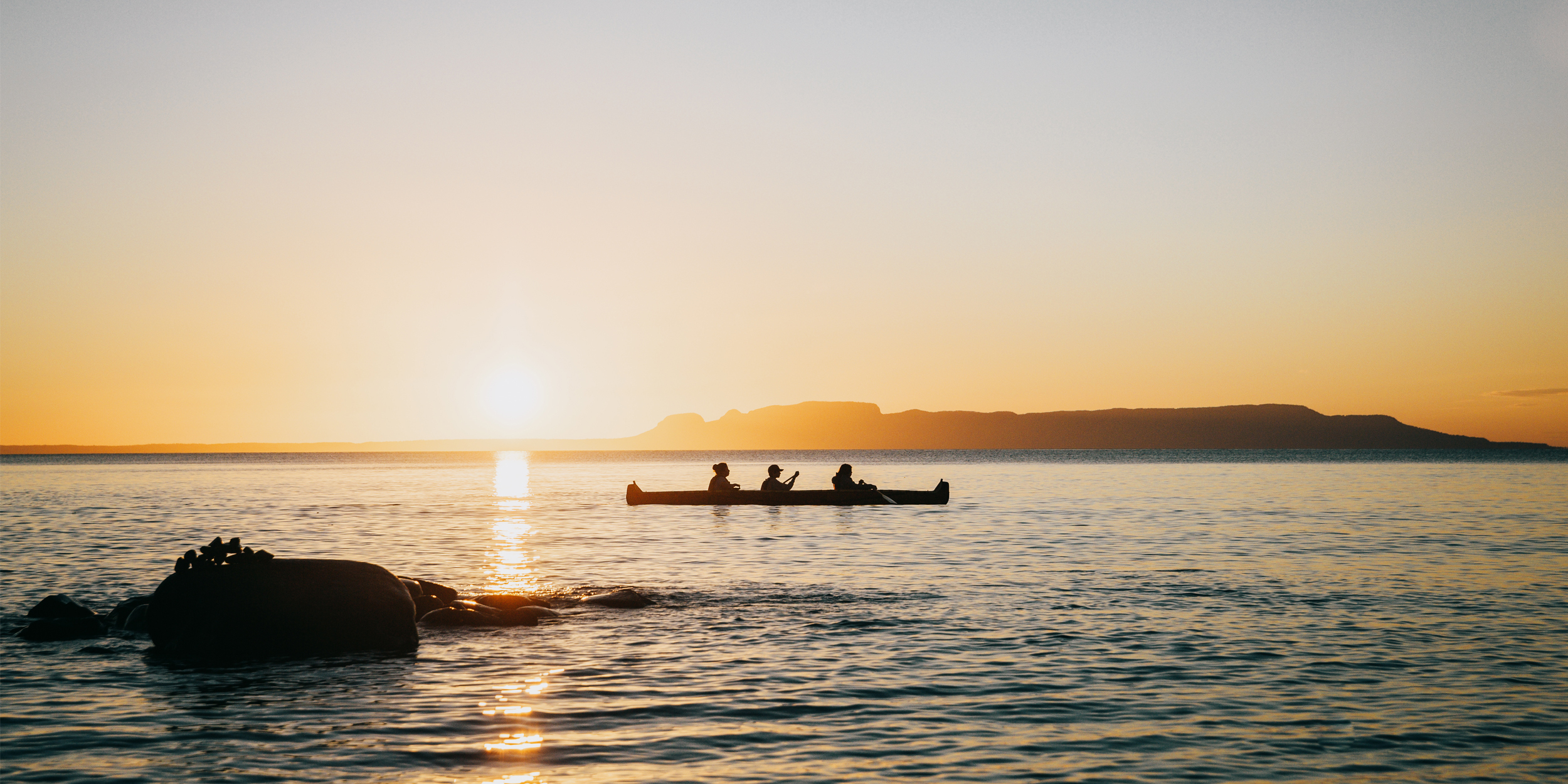 Trois personnes à bord d’un long canot pagayant au coucher du soleil sur les eaux bleues immobiles du lac Pass.