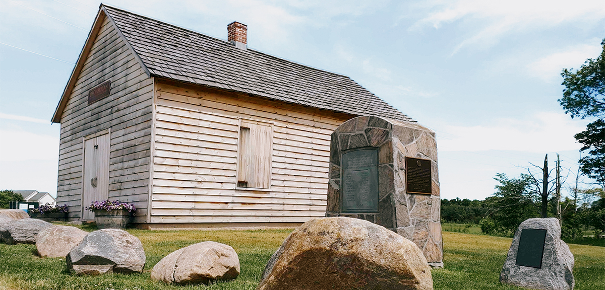 Kleine historische Holzkirche, umgeben von Steinmarkierungen und Gedenkstätten auf einem grasbewachsenen Gelände.