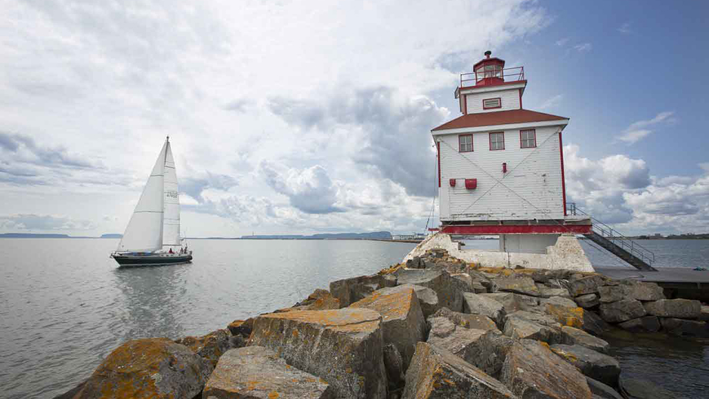 A sailboat sails past a white and red lighthouse atop a rocky coastline