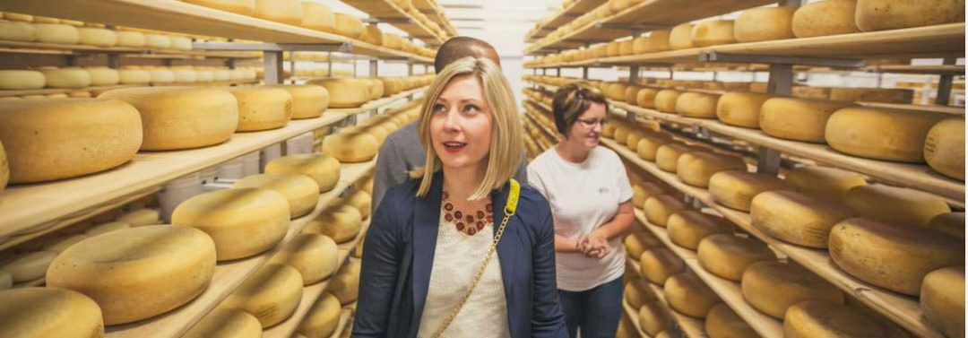 People taking a tour, walking through an isle with shelves full of large round cheese