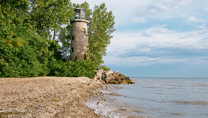 Cobblestone lighthouse surrounded by trees on the edge of a rocky shoreline.