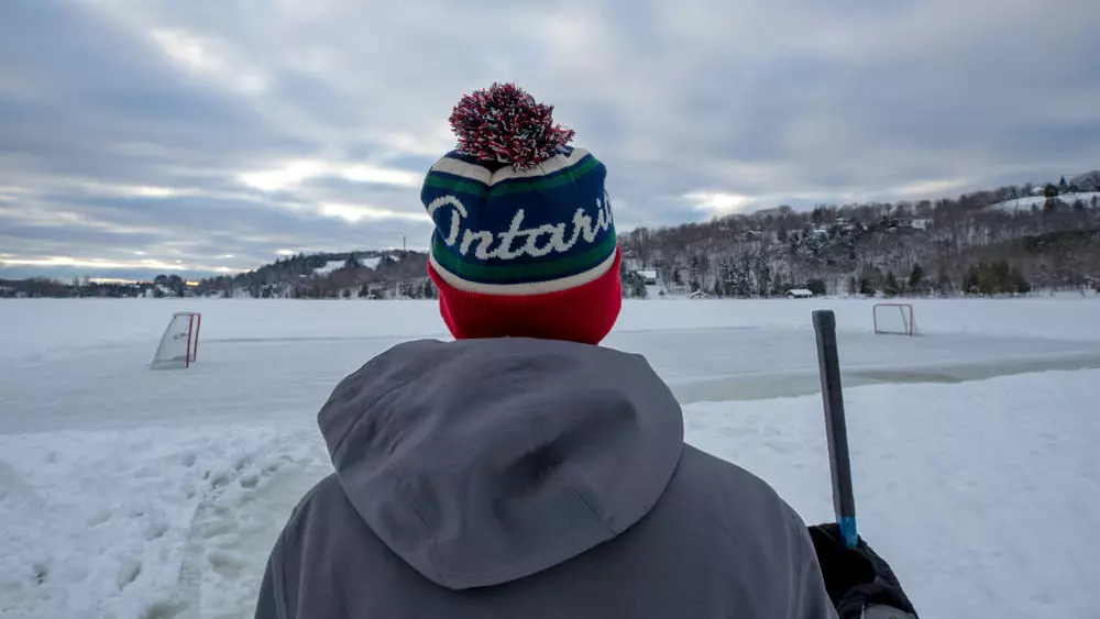 Un joven se encuentra frente a una pista de hockey sobre hielo en un estanque sosteniendo un palo de hockey.