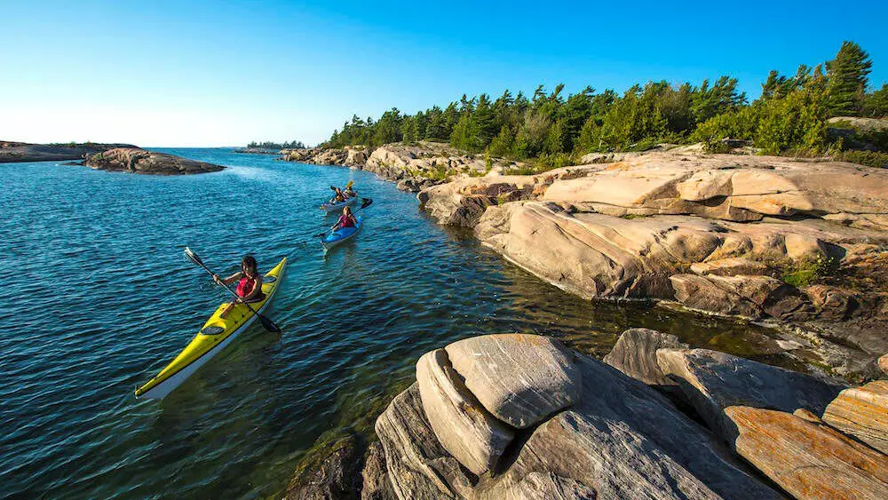 Three people paddling along the lake close to rocky land with trees.