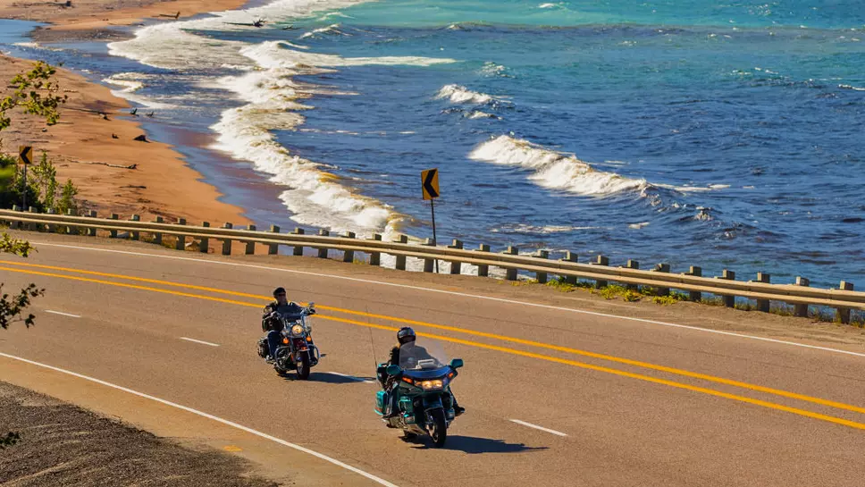 Two motorcyclists ride past a sandy beach with white capped waves crashing in.