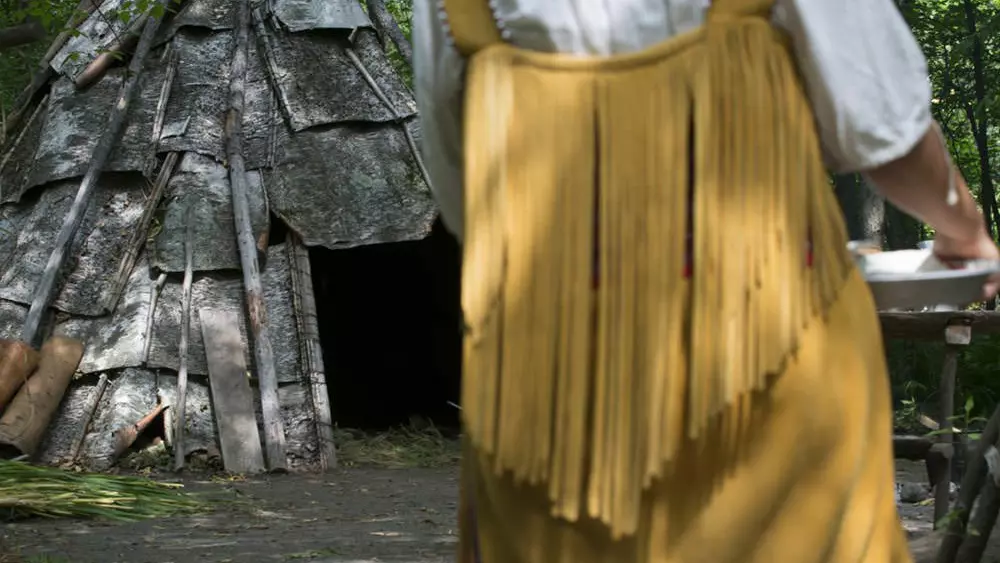 A woman carries a plate to a traditional Indigenous dwelling.