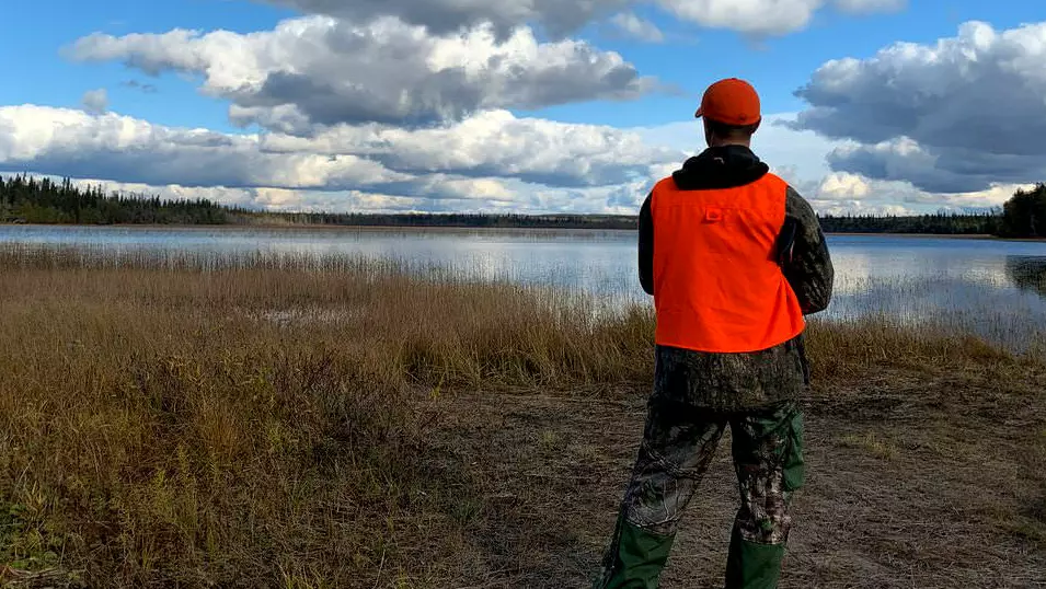 オレンジ色のハンティング キャップとベストを身に着けたハンターが湖を眺めています。