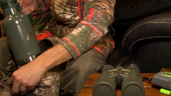 Un hombre vestido con camuflaje empaca binoculares, un termo y otros equipos de caza.