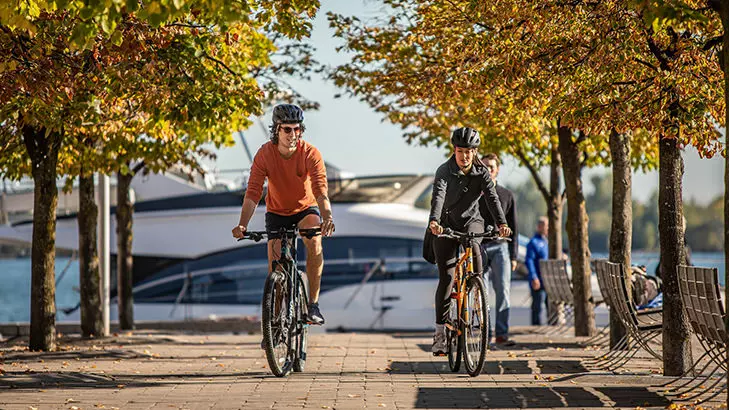 Deux personnes faisant du vélo le long d'arbres aux couleurs de l'automne avec un bateau assis en arrière-plan