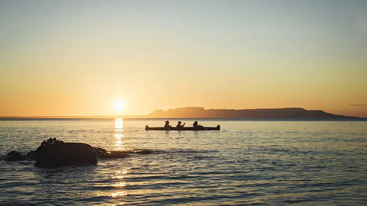 Tres personas remando en una canoa durante el atardecer con el Gigante Durmiente al fondo