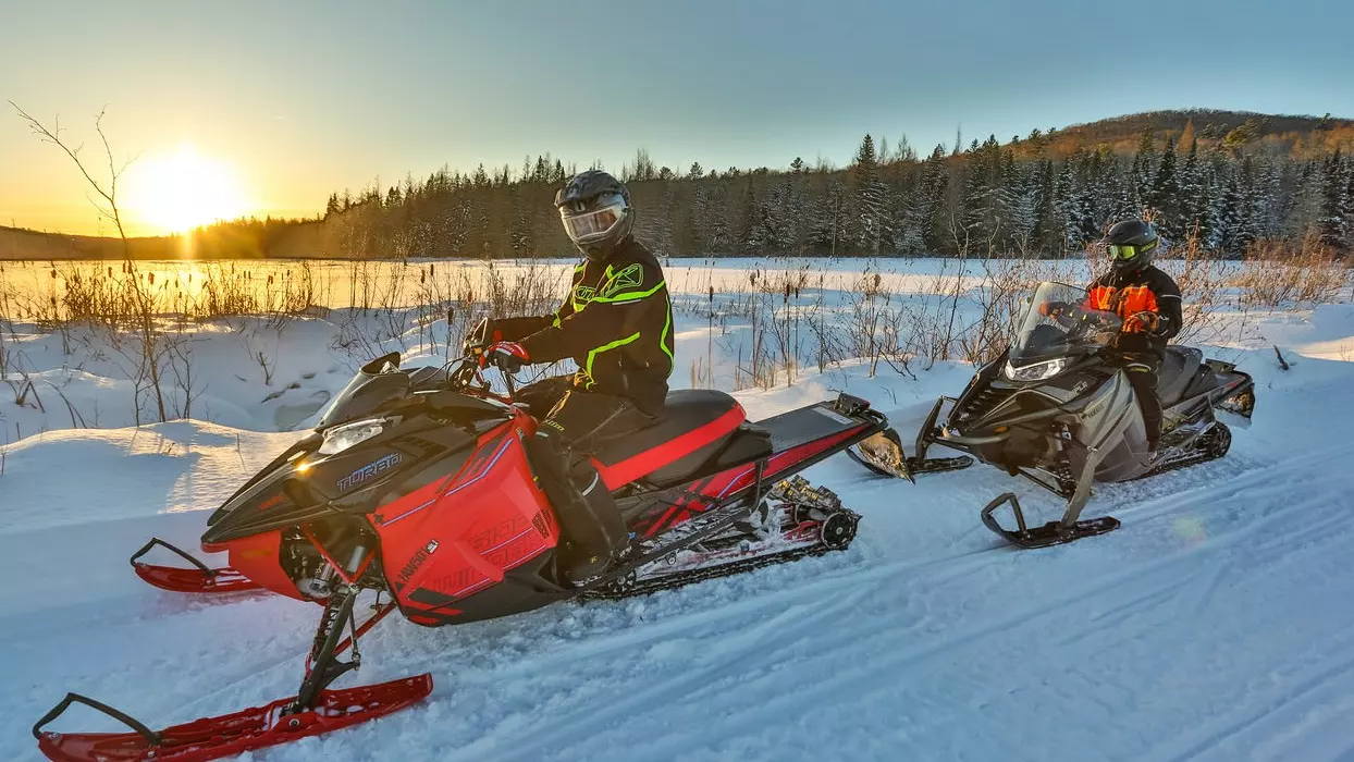 Two snowmobilers stop to watch the sunset over the snow.