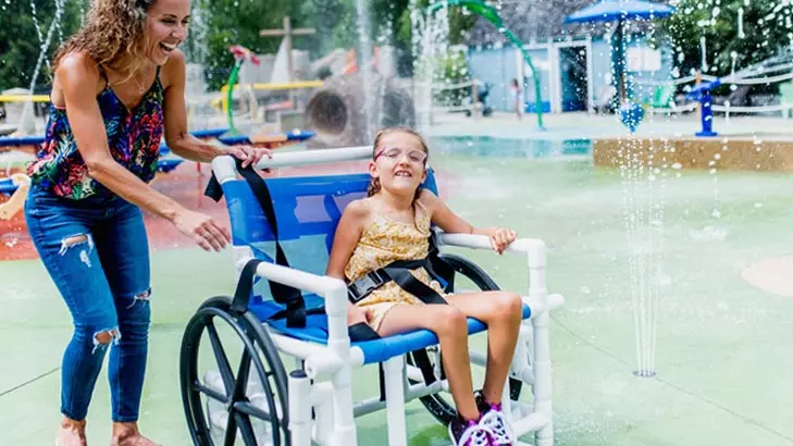 A woman and a young girl sitting in a wheelchair enjoy playing in a water fountain.