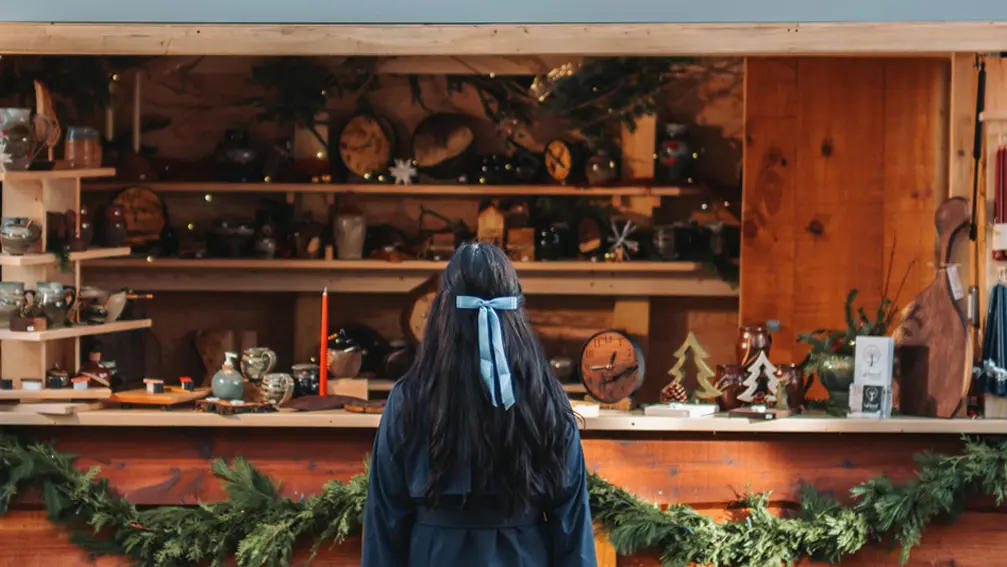 Jeune fille aux cheveux noirs foncés avec boucle bleue regardant vers un stand de marché des Fêtes où sont exposées des décorations en bois.