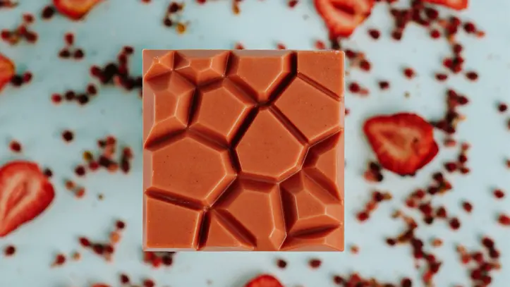 Ein einzigartiger geometrischer Erdbeerschokoladenriegel vor einem pastellfarbenen Hintergrund, bestreut mit getrockneten Erdbeeren und Beerenstücken.