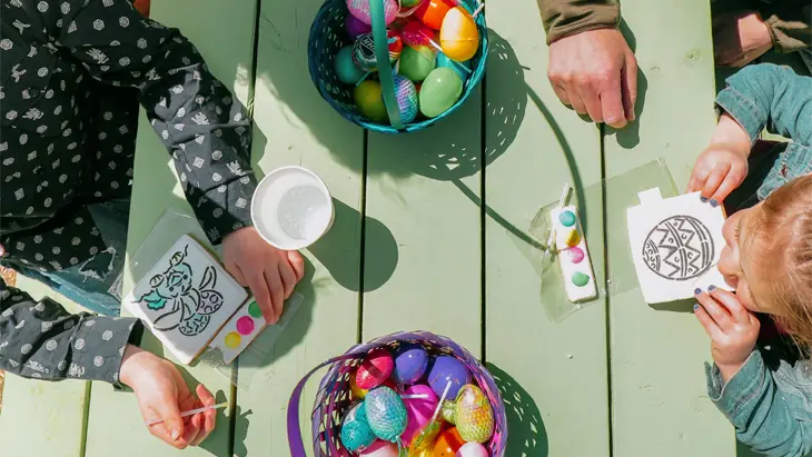 Una familia reunida alrededor de una mesa de picnic verde, decorando manualidades de Pascua y huevos coloridos bajo el sol.