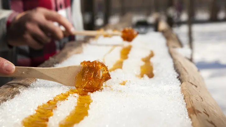 Primer plano de un caramelo de arce enrollado en palitos sobre la nieve en una choza de azúcar, con jarabe dorado brillando al sol.