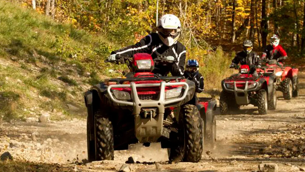 Eine Person mit einem weißen Helm fährt auf einem roten ATV und führt drei andere Personen auf ATVs eine unbefestigte Straße entlang.
