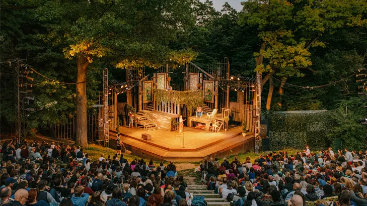 A crowd gathered at an outdoor amphitheatre surrounded by trees, watching a play unfold on a warmly lit stage at dusk.