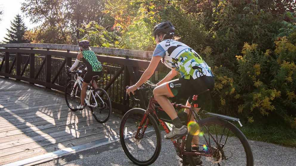 Two gravel bike cyclists cross a wooden bridge on a cycling trail near Collingwood.