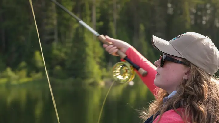 A woman casts using a fly fishing rod over a river on a summer day.