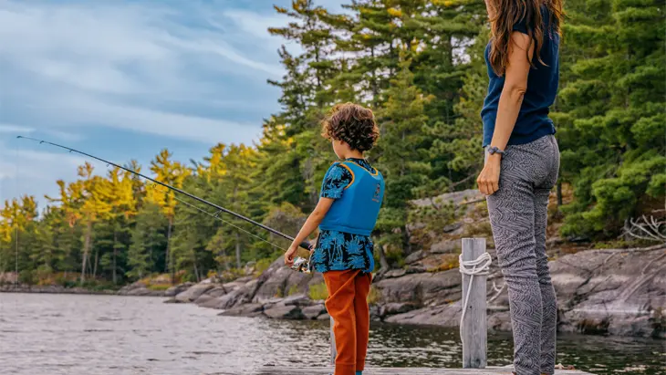 프랑스 강의 부두에서 어른과 함께 낚시를 하는 어린아이. 주변은 바위가 많은 해안과 푸른 나무로 둘러싸여 있습니다.