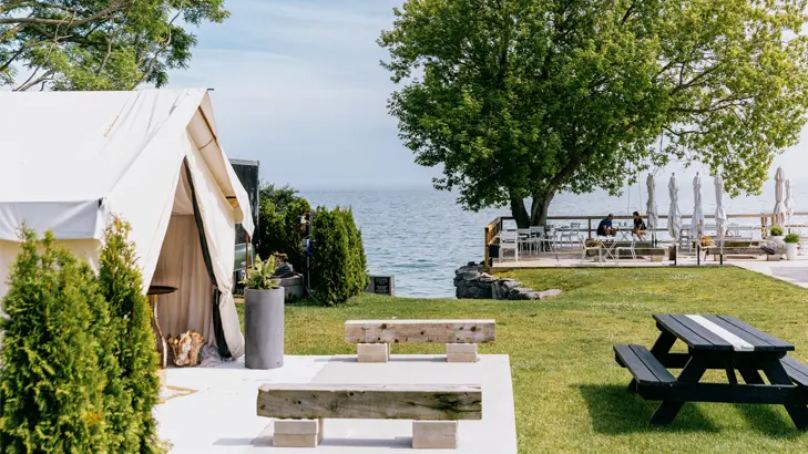 Ein Blick vom Lakeside Motel in Prince Edward County mit einem Glamping-Zelt und einer Terrasse am Seeufer, auf der Gäste unter einem Baum am Wasser sitzen.