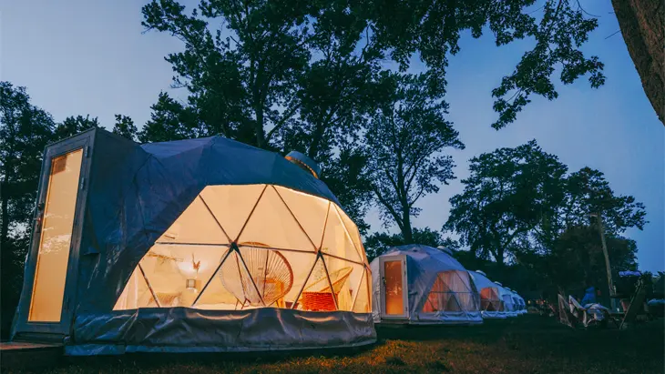 A row of illuminated glamping tents at Lungovita Beach Retreat in Windsor Essex, glowing under the night sky with trees and picnic benches nearby.