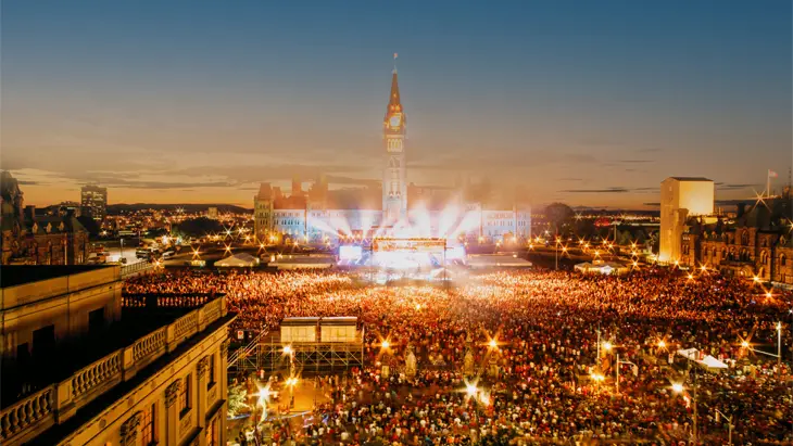 Des milliers de personnes devant la scène du spectacle de la fête du Canada sur la Colline du Parlement à Ottawa, éclairées de stroboscopes lumineux.