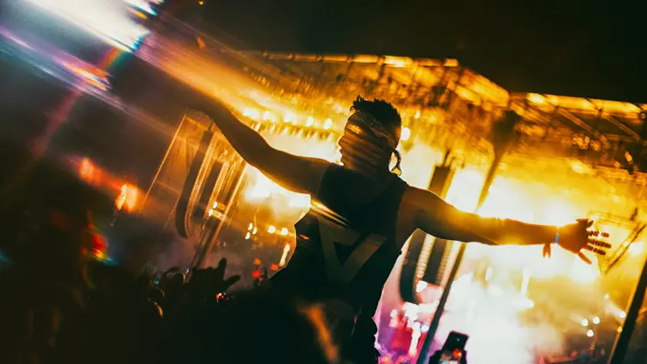 Ein Besucher des VELD Music Festival sitzt mit ausgestreckten Armen auf den Schultern einer Person und wird von den hellen Lichtern der Bühne angestrahlt.