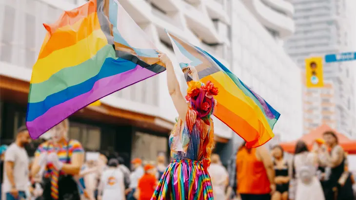 Ein Teilnehmer einer Pride-Parade in einem Regenbogen-Outfit schwenkt mitten auf einer belebten Kreuzung zwei Pride-Flaggen.