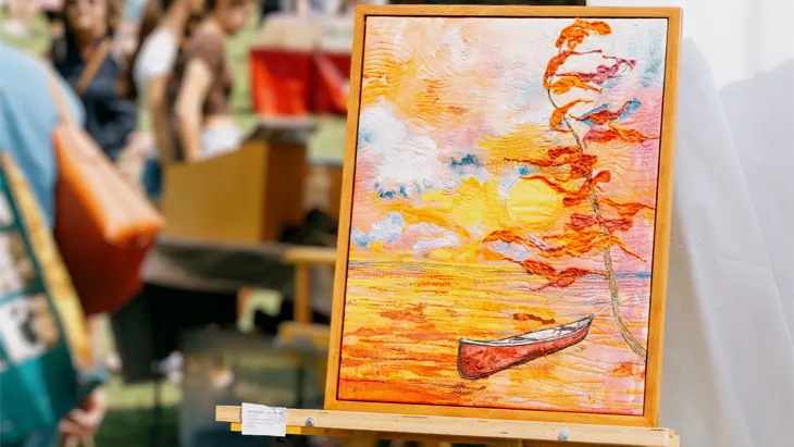 Toile représentant un canot rouge flottant sur l’eau sous un coucher de soleil orange, posée sur un chevalet à une exposition d’art estivale.
