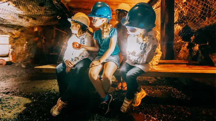 ダイナミック アースの岩壁の薄暗い地下洞窟の中で、ヘルメットをかぶった 4 人の子供たちが木製のベンチに座っています。