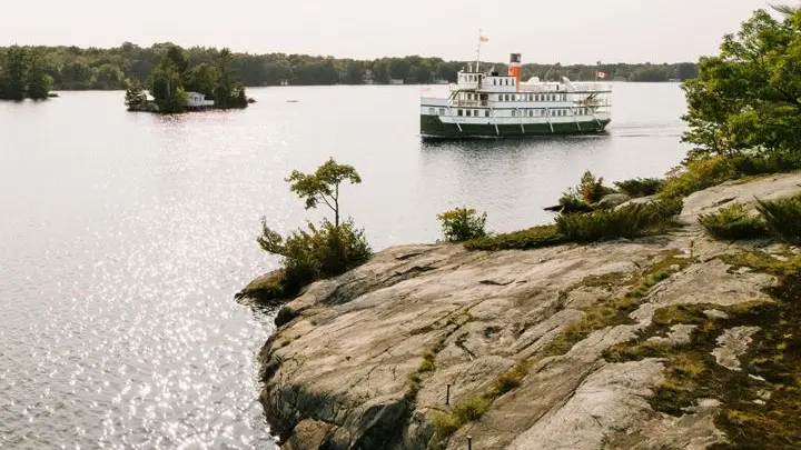 A heritage passenger steamship cruises past rocky shoreline and forested islands in Muskoka.