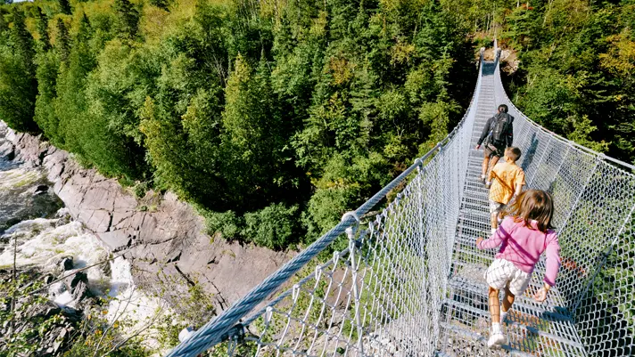 Una mujer y dos niños pequeños caminan por un puente colgante sobre rápidos en el Parque Nacional Pukaskwa en un día de verano.