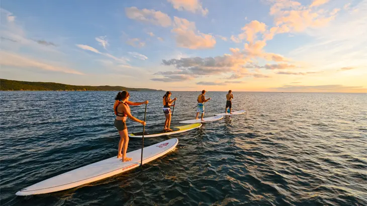 オンタリオ州ワワの静かな水面で夕日に向かってスタンドアップ・パドル・ボードを楽しむ4人の友人たち。