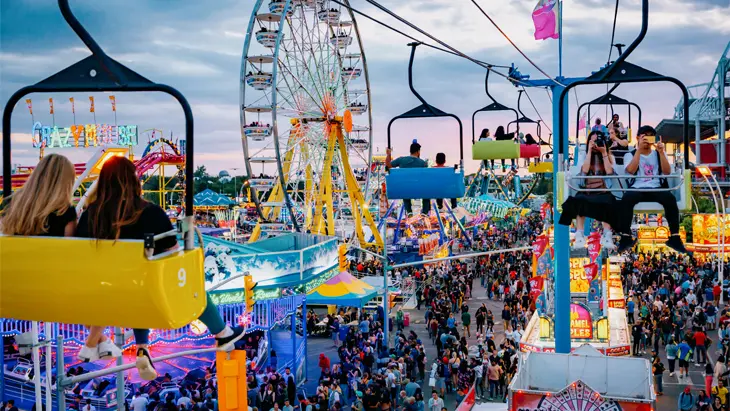 Bajo un cielo púrpura, un animado parque de atracciones, repleto de visitantes, presenta una gigantesca noria, atracciones coloridas y un telesilla en la Exposición Nacional Canadiense en Toronto.