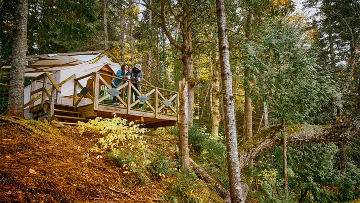 Deux personnes debout sur la terrasse d’une cabane en bois perchée sur une colline surplombant une forêt luxuriante à Bartlett Lodge, à Algonquin.