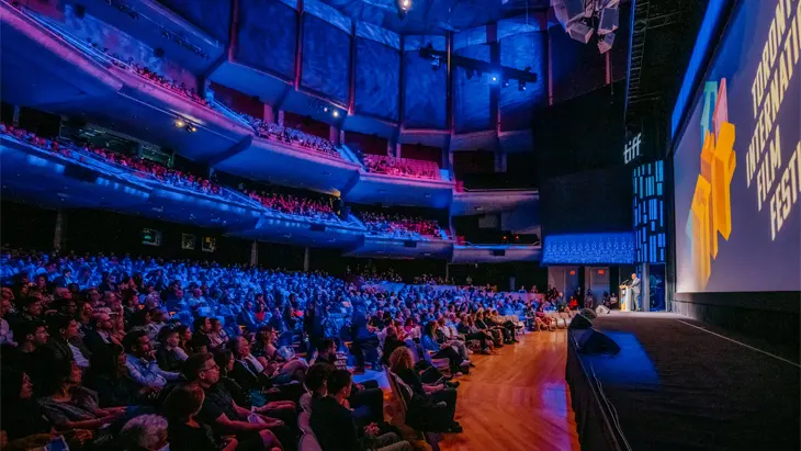 A large, full TIFF theatre lit in blue light, with participants looking towards a spotlighted speaker on stage.