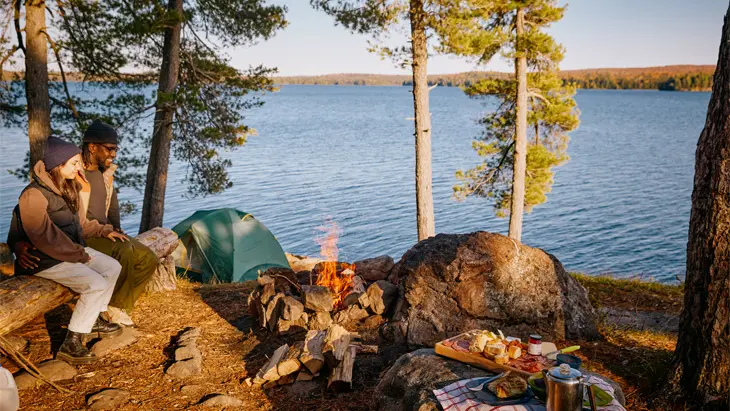 A couple sits on a log bench in front of a roaring fire on a camp site overlooking a vast lake.