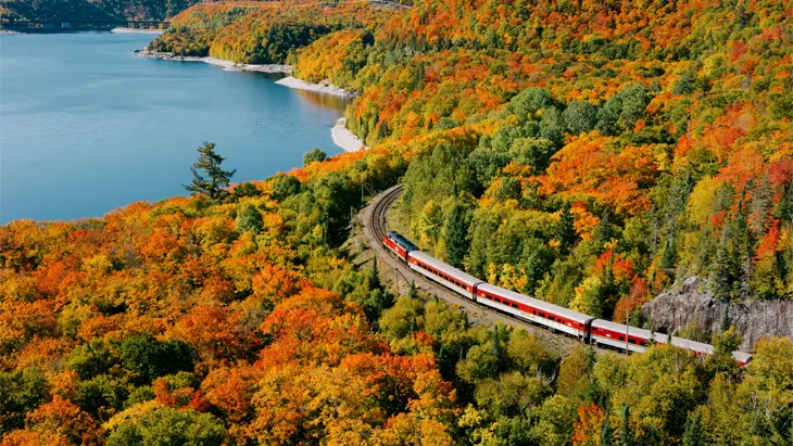Aerial view of a train travelling past a lake and forest during the peak of fall colour.