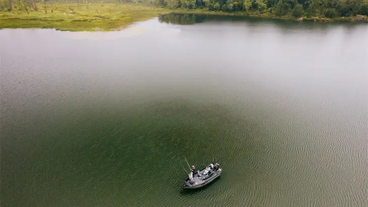 Aerial view of a fishing boat floating on a calm green lake with lush trees in the distance.