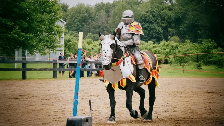 Un caballero montado en un caballo blindado vestido de rojo y amarillo frente a frondosos árboles y una audiencia.
