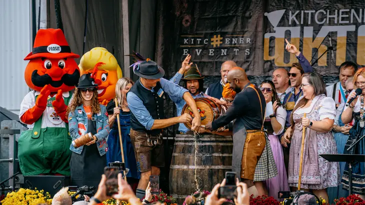 Eine Menge jubelt in Richtung einer Oktoberfestbühne, wo in Lederhosen gekleidete Künstler ein Fass Bier ausschenken.