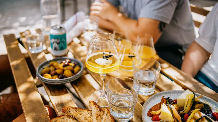 Escena de cena al aire libre en Toronto con gente reunida alrededor de una mesa de madera, disfrutando de bebidas y platos compartidos de pan, queso y aceitunas.