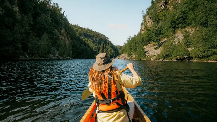 Vue de dos d’une femme portant un chapeau gris à bord d’un canot sur une rivière calme et sinueuse entre des collines bordées de grands arbres.