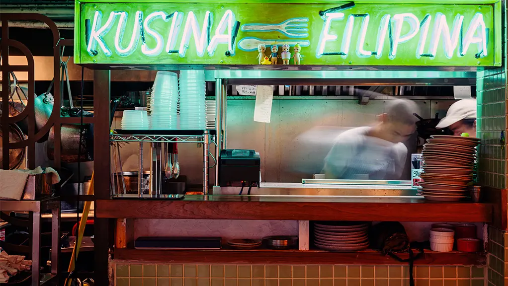 A chef prepares a dish at a food stall with a green neon sign, "Kusina Filipina." Stacks of plates and utensils are in view.