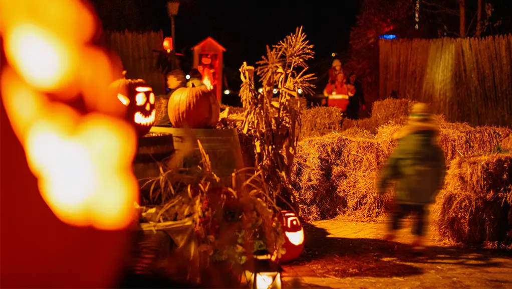 A child walks through a spooky Halloween trail with hay bales and illuminated pumpkins lighting the path.