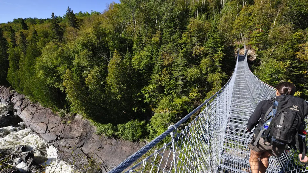 Trois personnes avec de gros sacs à dos en randonnée dans le parc national Pukaskwa, traversant un terrain rocailleux surplombant une eau bleue.